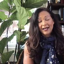 A woman with long dark hair is speaking in an indoor setting about women in politics. She is wearing a navy blue scarf with white patterns over a black sleeveless top. Behind her are large leafy plants and a bookshelf filled with various books. The PBS NewsHour logo is in the bottom left corner.