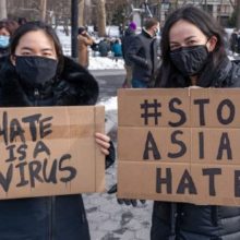 Two women in winter coats and face masks at a Stop Asian Hate rally, holding up signs.