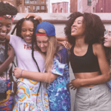 A group of five diverse young women stand close together, smiling and laughing. Dressed in colorful, casual clothing, they appear to be enjoying each other's company. The urban backdrop with buildings and a mural adds charm. They resemble the vibrant characters from HBO's "Betty.