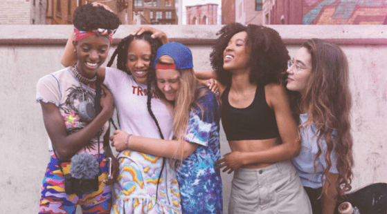 A group of five diverse young women stand close together, smiling and laughing. Dressed in colorful, casual clothing, they appear to be enjoying each other's company. The urban backdrop with buildings and a mural adds charm. They resemble the vibrant characters from HBO's "Betty.