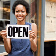 A smiling person stands outside a store, holding up a black and white "OPEN" sign. They are wearing a sleeveless blue top, promoting the movement to support Black businesses. Behind them is a window reflecting the street and menus posted on the wall.