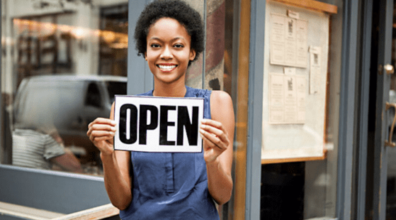 A smiling person stands outside a store, holding up a black and white "OPEN" sign. They are wearing a sleeveless blue top, promoting the movement to support Black businesses. Behind them is a window reflecting the street and menus posted on the wall.