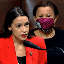 A woman in a bright red blazer is speaking passionately at a podium, with a microphone in front of her. Behind her, another woman wearing black and a red and purple patterned mask listens attentively. Both are seated in brown leather seats, as if discussing key examples from 2020.