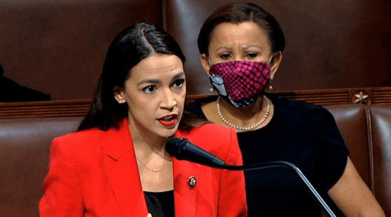 A woman in a bright red blazer is speaking passionately at a podium, with a microphone in front of her. Behind her, another woman wearing black and a red and purple patterned mask listens attentively. Both are seated in brown leather seats, as if discussing key examples from 2020.