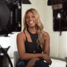 A person with long blonde hair is sitting and smiling, surrounded by camera equipment. The individual is wearing a black sleeveless top and dark pants in what appears to be a professional studio, perhaps prepping for a Netflix documentary on trans representation and disclosure.