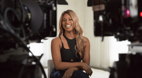 A person with long blonde hair is sitting and smiling, surrounded by camera equipment. The individual is wearing a black sleeveless top and dark pants in what appears to be a professional studio, perhaps prepping for a Netflix documentary on trans representation and disclosure.