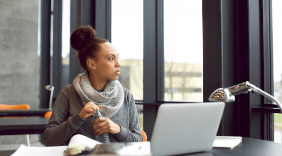 A person with their hair in a bun sits at a desk with a laptop, notebook, and desk lamp. They are holding a water bottle and looking out the window in front of them, perhaps contemplating their next post for one of the BLACK AT Instagram accounts. The background outside the window appears to be an urban setting.