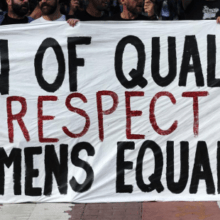 A group of people holds up a large white banner with bold black and red letters reading "MEN OF QUALITY RESPECT WOMEN'S EQUALITY" during a public demonstration, showcasing how to be a true male ally.