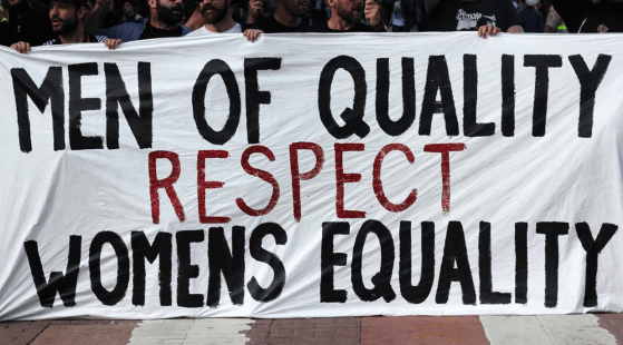 A group of people holds up a large white banner with bold black and red letters reading "MEN OF QUALITY RESPECT WOMEN'S EQUALITY" during a public demonstration, showcasing how to be a true male ally.