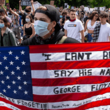 A large crowd of protesters marches in the street. A person at the front holds an American flag with messages like "I CAN'T BREATHE," "SAY HIS NAME," and "GEORGE FLOYD." Many participants wear masks, and signs are visible in the background, reflecting the intense turbulence of 2020 within American society.