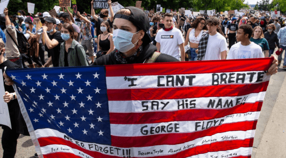 A large crowd of protesters marches in the street. A person at the front holds an American flag with messages like "I CAN'T BREATHE," "SAY HIS NAME," and "GEORGE FLOYD." Many participants wear masks, and signs are visible in the background, reflecting the intense turbulence of 2020 within American society.