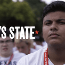 A close-up of a young man with a serious expression, wearing a red lanyard that reads "Texas Boys State." He is part of a group of similarly dressed individuals, blurred in the background. Text on the image reads "Boys State: Living In It" with a star on each side and the Apple TV+ logo.