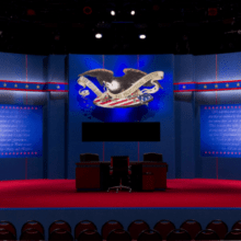 A debate stage with blue walls decorated with red, white, and blue stripes and stars, featuring a large eagle emblem above a screen. Two wooden podiums for the presidential debates are center stage on a red carpet. Rows of empty chairs, awaiting an audience eager to discuss political topics, are visible in the foreground.