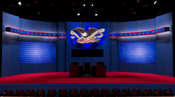 A debate stage with blue walls decorated with red, white, and blue stripes and stars, featuring a large eagle emblem above a screen. Two wooden podiums for the presidential debates are center stage on a red carpet. Rows of empty chairs, awaiting an audience eager to discuss political topics, are visible in the foreground.