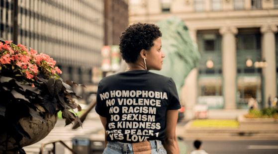 A person stands in an urban setting wearing a black t-shirt with white text that reads "No Homophobia, No Violence, No Racism, No Sexism, Yes Kindness, Yes Peace, Yes Love." This embodiment of values and inclusivity is set against a backdrop of a street with tall buildings and a potted plant with flowers.