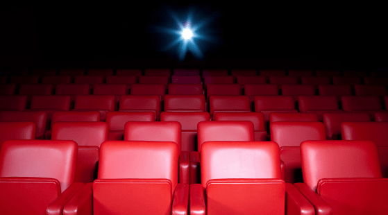 Empty red theater seats face a brightly lit screen in a dark cinema. The glow from the screen illuminates the rows, creating a contrast between the vibrant red seats and the dim surroundings, awaiting the keen eye of a female film critic to breathe life into its narrative.