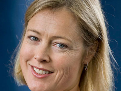 A woman with shoulder-length blonde hair and blue eyes is smiling. She is wearing small earrings and is photographed against a blue background, where it almost seems like her cheerful expression defies the notion that life can be an uphill battle.