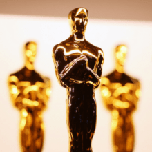 A row of shiny, golden Oscar statuettes stands against a blurred background. The awards, representing the pinnacle of the film industry, depict a stylized knight holding a sword. Closely lined up, they create a pattern of reflections on their polished surfaces.