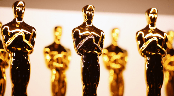 A row of shiny, golden Oscar statuettes stands against a blurred background. The awards, representing the pinnacle of the film industry, depict a stylized knight holding a sword. Closely lined up, they create a pattern of reflections on their polished surfaces.