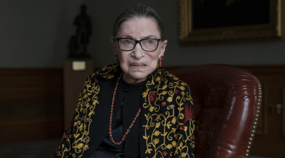 An elderly woman with short, pulled-back hair and glasses is sitting on a leather chair. She is wearing a black outfit with a decorative, embroidered shawl and red jewelry. The background features a dark statue and a framed portrait of RBG, portraying her as an inspiring role model for women everywhere.