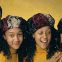 A family of four poses against a yellow backdrop. The man on the left wears a suit, the woman on the right wears dark clothing, and the two twin girls in the middle wear matching yellow tops and velvet hats with floral patterns. All are smiling and looking at the camera, reminiscent of beloved Black sitcoms.