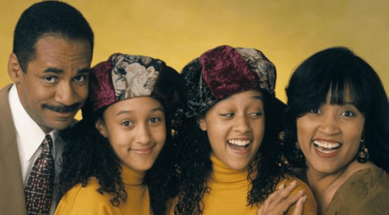 A family of four poses against a yellow backdrop. The man on the left wears a suit, the woman on the right wears dark clothing, and the two twin girls in the middle wear matching yellow tops and velvet hats with floral patterns. All are smiling and looking at the camera, reminiscent of beloved Black sitcoms.