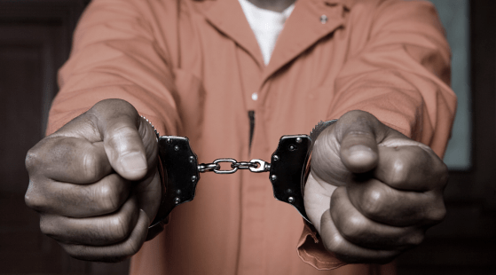 A black person in an orange jumpsuit holding out their wrists, which are handcuffed.