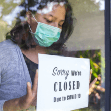 A picture of a woman in a face mask putting a sign in a store window. The sign says "Sorry, we're closed due to COVID-19"