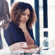 Two women in a corporate setting, looking at documents and a computer.