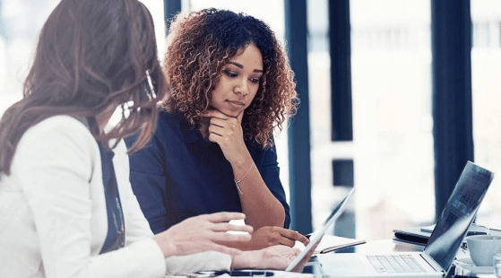 Two women in a corporate setting, looking at documents and a computer.