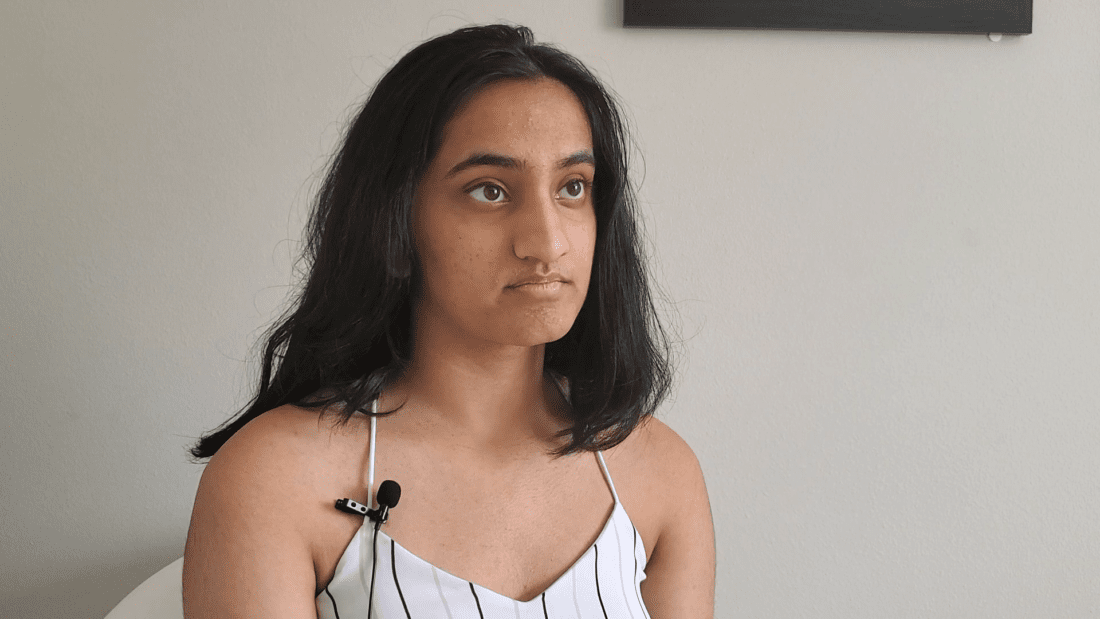 A person with long dark hair is seated in front of a light-colored wall, wearing a white top with black stripes. They are looking slightly off-camera with a neutral expression, and there is a small microphone clipped to their top, giving the impression of being part of a documentary on representation.