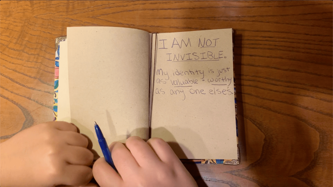 A pair of hands holding a pen and writing in a notebook. The notebook has a message written on the right page that reads, "I am not invisible: My identity is just as valuable & worthy as anyone else's." The left page is blank. This scene captures powerful representation resting on a wooden table.