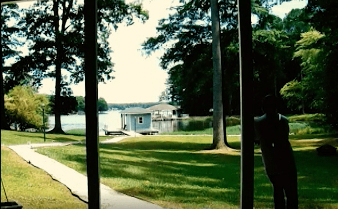 A tranquil lakeside scene viewed from a porch, showing a grassy lawn leading to a dock with a small boathouse. Tall trees surround the water, and a shadowy figure stands in the foreground, partially obscured by the porch supports. It feels almost like stepping into a still frame from an artful documentary.