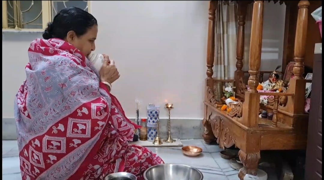 A person in a red and white shawl sits on the floor near a small wooden shrine, performing a prayer ritual. They are blowing into a conch shell, with candles, flowers, and offerings arranged around the shrine in a well-lit room—a serene representation captured as if for a documentary.