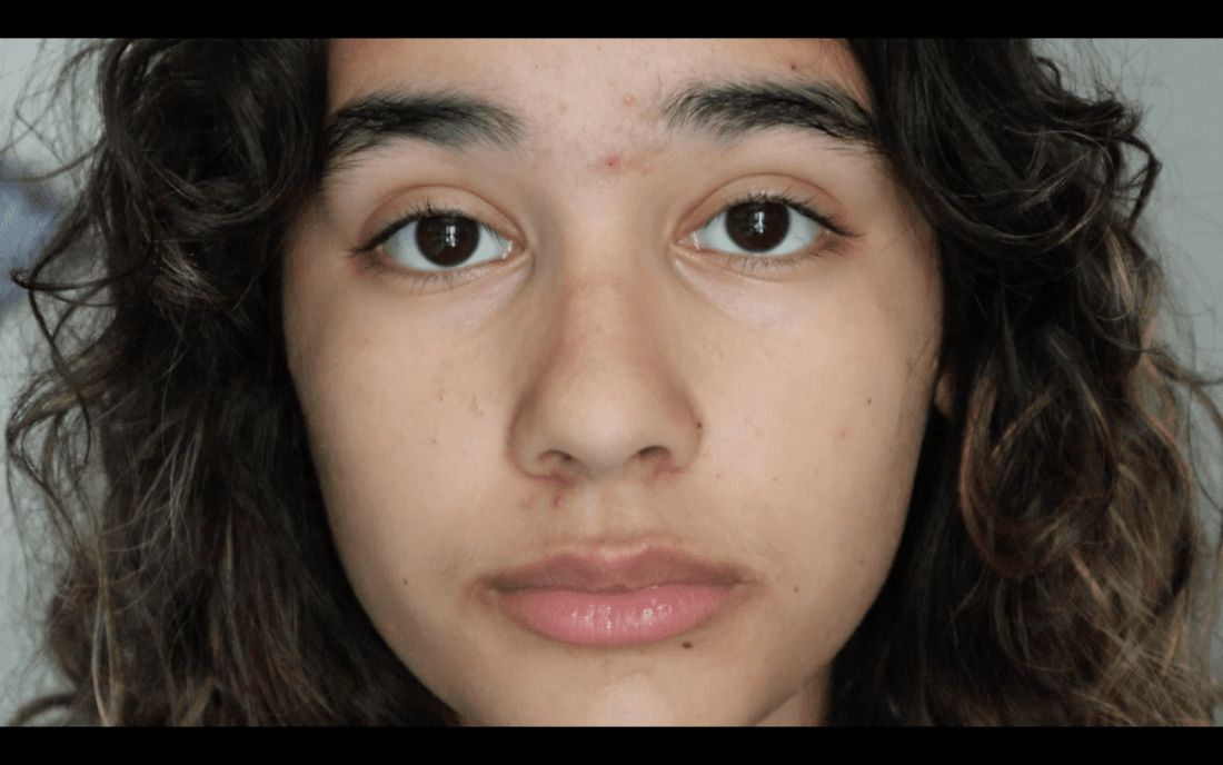 A close-up photo of a biracial person with long, curly hair and brown eyes, gazing directly into the camera. The person's facial expression appears neutral, and the background is out of focus, giving it a documentary feel.