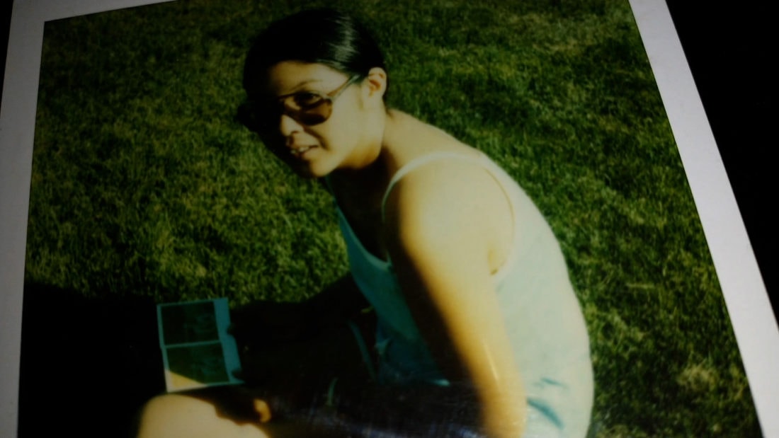 A person wearing sunglasses and a sleeveless top sits on the grass, looking towards the camera. They have short dark hair pulled back and hold an open book on their lap. It's a sunny outdoor setting, reminiscent of my-grandma-fighter's pose from the old family documentary.