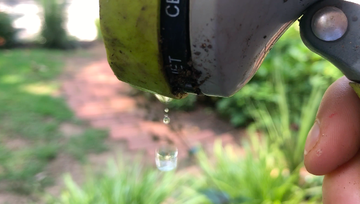 Close-up of a garden hose nozzle leaking water droplets. The background is a garden with green plants and a red-brick path, blurred to focus on the water droplets. The nozzle shows signs of dirt and wear, almost as if telling its own documentary story of everyday use and resilience.