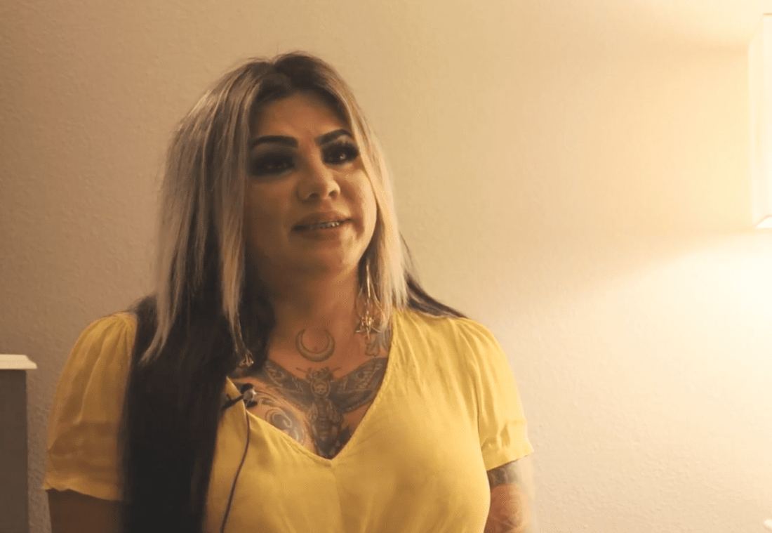 A woman with long, dark hair featuring lighter highlights, wearing a yellow shirt, is shown in a softly lit indoor setting. She has visible tattoos on her chest and is speaking into a microphone attached to her shirt, embodying representation and self-love in this compelling documentary.