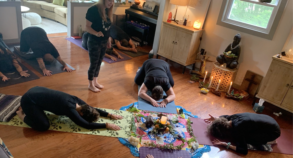 A small group of people participate in a healing yoga session inside a cozy room with wooden floors. They are in various child's poses around a colorful mandala arrangement in the center. A female instructor stands and guides them. The room is softly lit with candles and lamps, creating a reiki-infused ambiance.