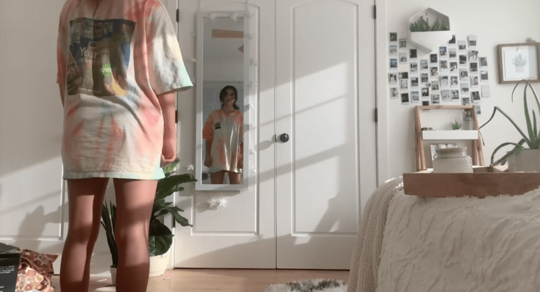 A person stands in a brightly lit bedroom wearing a colorful tie-dye T-shirt, facing a full-length mirror attached to white closet doors. The room features plants, a bed with a white blanket, and a wall decorated with photos and small frames, reflecting the moments captured in their personal documentary.