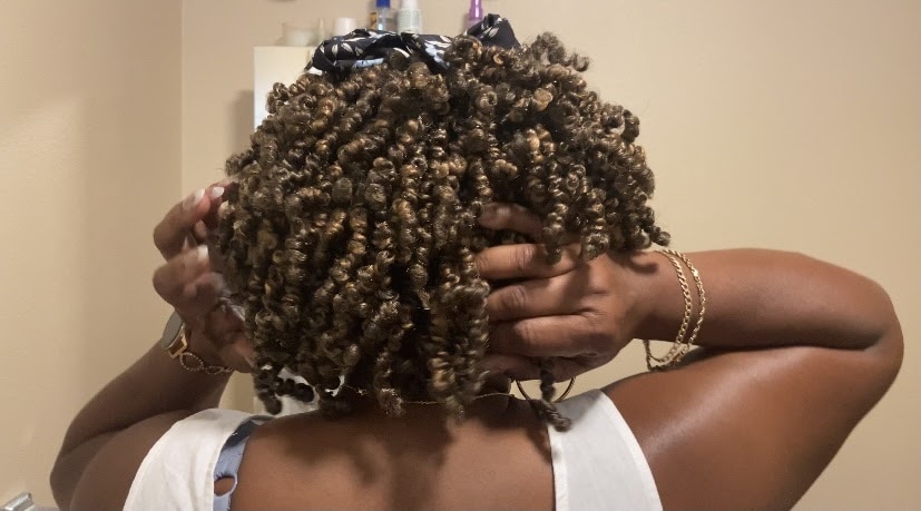 A person with curly, shoulder-length hair stands with their back to the camera, using both hands to style or adjust their hair. They are wearing a white top and a few gold bracelets on their right wrist. Various hair care products are visible in the background, reminiscent of a grandmother's loving touch.