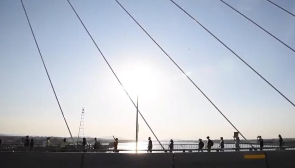 A group of people walks across a cable-stayed bridge during sunset, with the sun visible and reflecting off the water in the background. The scene, reminiscent of a poignant documentary, features prominently the cables creating diagonal lines across the sky.