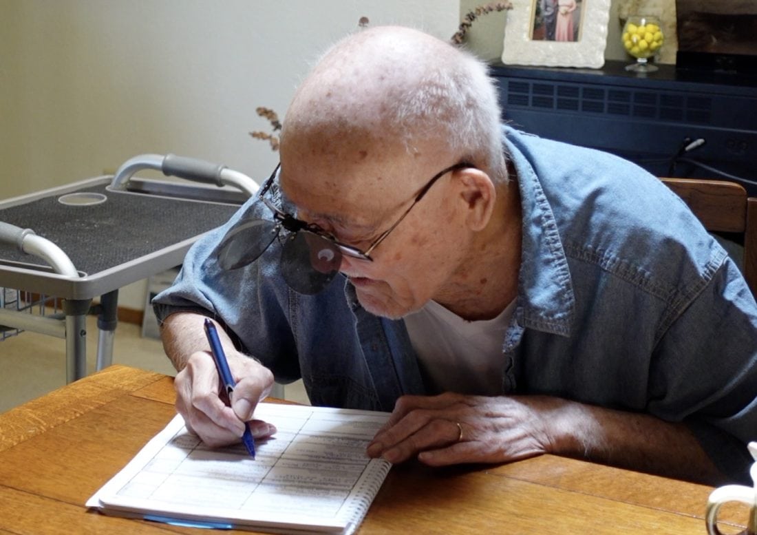 Papa, an elderly man with a bald head and glasses clipped with magnifying lenses, writes in a notebook on the table. Wearing a denim shirt and white t-shirt, he sits peacefully. In the background, his walker and a table with decorative items subtly hint at the care he receives for dementia.