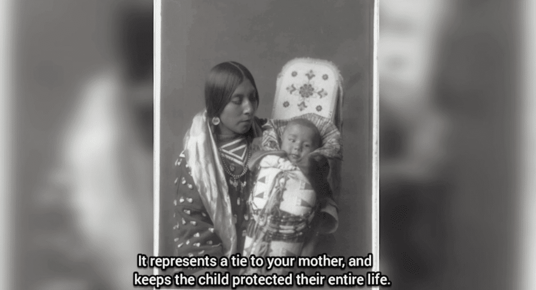 A vintage photograph shows a woman dressed in traditional attire holding a baby in a decorated cradleboard. The caption reads, "It represents a tie to your mother, like an unbroken cord, and keeps the child protected their entire life.