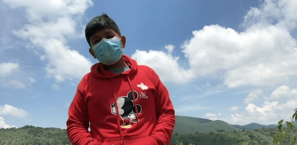 A young person stands outdoors wearing a red hoodie with a Mickey Mouse design and a blue face mask. Behind them, there is a scenic view of green hills under a partly cloudy blue sky, reminiscent of scenes from a heartfelt documentary asking, "why-do-you-take-away-our-happiness?