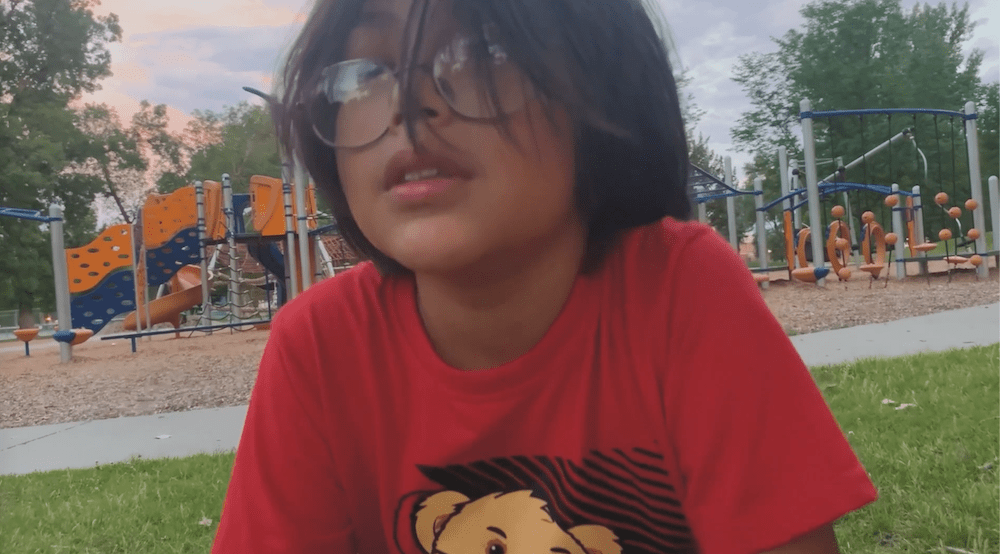 A young child with shoulder-length dark hair and glasses is sitting on the grass in front of a playground. Wearing a red T-shirt with a bear design, they smile softly. Trees and playground equipment dot the background, evoking the scenes often captured in heartfelt documentaries about indigenous communities.