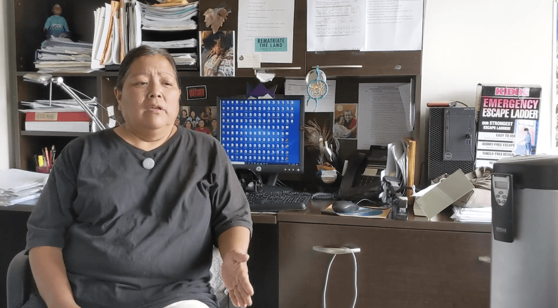 A person wearing a black shirt is seated in an office with various papers and objects on the desk and shelves behind them. A computer screen displays a blue background with white icons. A sign reading "Emergency Escape Ladder" is visible in the background, suggesting they're working on a documentary about the importance of cultural identity.