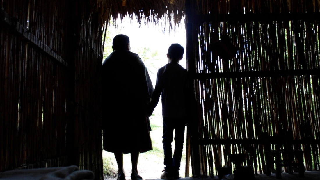 Two people are silhouetted against the light as they stand holding hands at the entrance of a rustic, thatched-roof hut. The surroundings outside appear bright, in contrast with the dim interior. The scene gives a sense of companionship and departure, like a sprouting journey documented in silence.