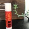 A 4-Pack Chapstick, labeled with "#RESPECT HER GAME" in bold red and yellow text, stands upright on a dark surface. In the background, there is a small green succulent plant alongside a partial view of a wooden object.