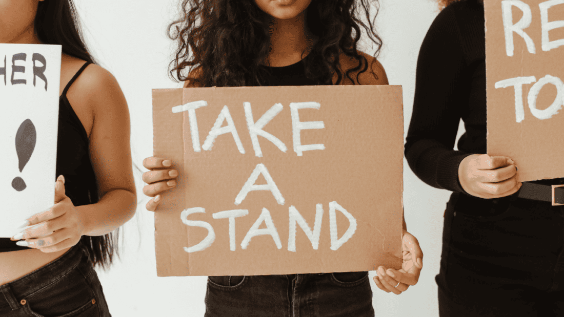 Three individuals are holding protest signs. One sign, held by a person in the center, reads "TAKE A STAND". The individuals are dressed in black, and the plain white background emphasizes the seriousness of their cause. Only their torsos and hands are visible, making their stance on abortion clear yet powerful.